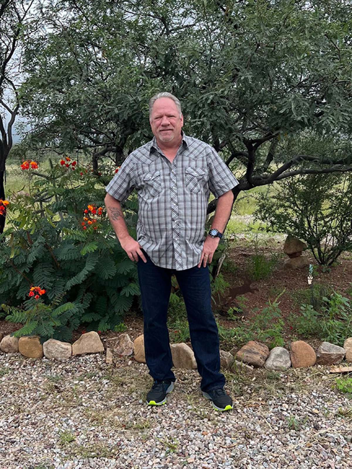 Larry Tait standing outdoors on a gravel path in front of a tree with his hands on his hips