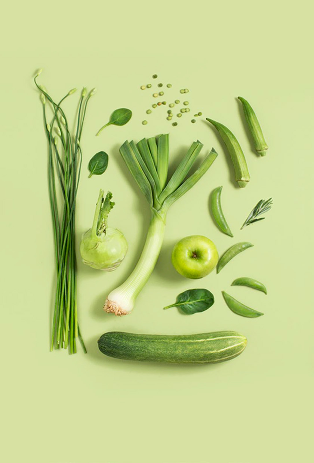 Green vegetables, fruit and herbs arranged on flat surface; scallions, leek, onion, spinach, cucumber, peas, okra, apple, rosemary and more